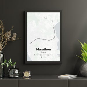 Op de afbeelding: Een ingelijste print met een kaart van een marathonroute in Athene. De zwarte lijn volgt de route tegen een witte en grijze achtergrond. Het woord "Marathon" staat vetgedrukt, met "Athene" eronder. De print bevat iconen en de afstand van 42,195 km.