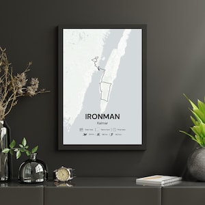 Op de afbeelding: Een ingelijste print met een zwarte rand, met een kaartomtrek van Kalmar, Zweden, met het label "IRONMAN Kalmar". De print bevat pictogrammen en afstanden: 3,8 km zwemmen, 180 km fietsen en 42,2 km hardlopen. De achtergrond is lichtgrijs.