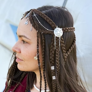 May include: A silver-toned hair accessory featuring delicate chains, small charms, and a circular filigree medallion. The piece is integrated into brown braided hair, with additional silver rings and beads attached to the individual braids.
