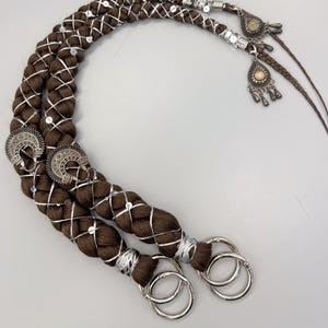 Puede incluir: Dos extensiones de cabello trenzado marrón con detalles plateados y dijes decorativos de metal. Las trenzas están adornadas con hilo plateado y pequeñas lentejuelas. Cada trenza tiene un gran anillo plateado en el extremo.