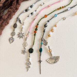 Op de afbeelding: Een verzameling kleurrijke dreadlock kralen en bedels. De kralen zijn geregen op verschillende gekleurde koorden, met een verscheidenheid aan edelstenen en decoratieve bedels, waaronder vleugels, zonnen, bloemen en een zwaard. Dreadlock accessoires.