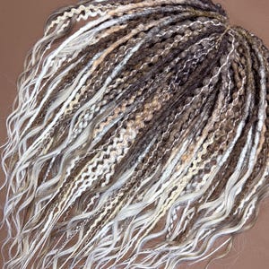 Puede incluir: Un conjunto de rastas sintéticas en tonos marrón, beige y blanco. Las rastas tienen una mezcla de texturas, incluyendo secciones lisas y onduladas. Los extremos de las rastas son sueltos y ondulados, creando un aspecto natural.