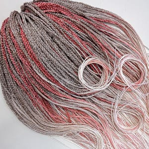 May include: A bundle of braided hair extensions in shades of gray, red, and light pink. The braids are tightly woven and arranged in a cascading pattern. Several strands of white and pink thread are coiled at the bottom.