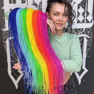 May include: A vibrant rainbow of braided hair extensions, featuring shades of blue, green, yellow, orange, and pink. The extensions are held up, showcasing the full spectrum of colors. The hair is styled in long braids.