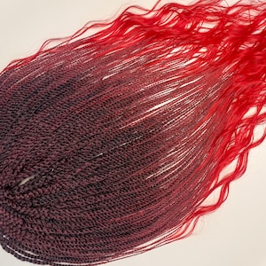 May include: A close-up of red and burgundy-colored hair extensions. The extensions are made of small, tightly woven braids that transition from a deep burgundy to a vibrant red. The ends of the hair have a wavy texture.