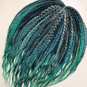 May include: A collection of synthetic dreadlocks in teal, turquoise, and grey hues. The dreads are a mix of braided and loose strands, creating a textured appearance. The ends of the dreadlocks have a wavy, flowing style.