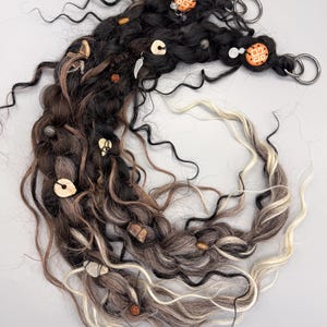 May include: A handcrafted hair wrap featuring a blend of black, brown, and blonde synthetic hair. Adorned with wooden beads, decorative charms, and orange patterned accents. Includes silver metal rings for attachment.
