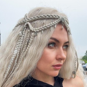 May include: A silver-toned hair accessory featuring delicate chains and small metal charms woven into platinum blonde braided hair. The headpiece includes multiple dangling pendants and metallic rings attached to the braids, creating a layered look.