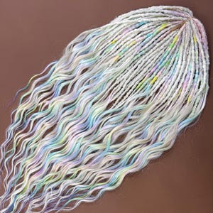 May include: A collection of handmade, wavy dreadlocks in shades of white, gray, and pastel colors. The dreads feature streaks of blue, green, yellow, and purple, creating a unique, colorful look. The dreadlocks are arranged on a brown surface.