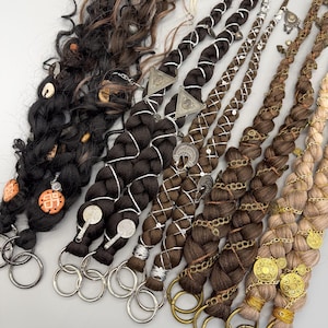 May include: An assortment of braided hair extensions in various shades of brown, black, and blonde. Each braid is adorned with decorative metal charms, beads, and silver or gold accents. The extensions are attached to large metal rings.