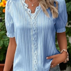 May include: Light blue blouse with short sleeves and a V-neckline. The blouse features vertical pleats and white lace trim along the neckline and center. A gold necklace with a star pendant is visible. The blouse is a casual top.
