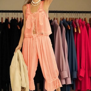 May include: A peach-colored pajama set with a tank top and cropped pants, trimmed with lace. The top has a tie closure. A beige jacket is held in the hand. Clothing hangs on a rack in the background.