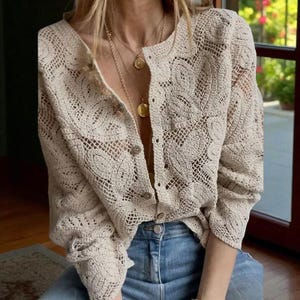May include: A cream-coloured, open-weave crochet cardigan with long sleeves and button fastenings. The intricate pattern features floral-like designs. The cardigan is paired with blue jeans and gold necklaces.