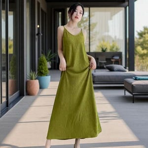 Peut inclure: Longue robe vert olive à fines bretelles et col en V. La robe présente une texture côtelée verticale. Le modèle se tient sur un patio en béton. La robe mesure environ 140 cm de long.