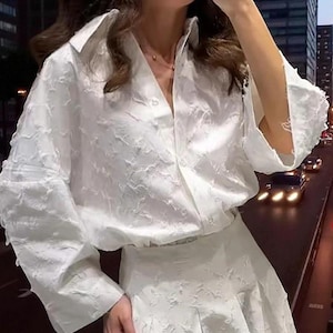 Könnte beinhalten: Ein weißes, strukturiertes Blusen- und Rock-Set. Die langärmlige Bluse hat einen Kragen und eine Knopfleiste. Der Rock ist plissiert. Das Outfit besteht aus einem Stoff mit einem subtilen, erhabenen Muster. Im Hintergrund ist eine Stadtstraße bei Dämmerung zu sehen.