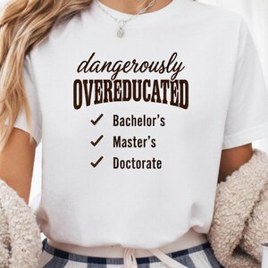 Könnte beinhalten: Weißes T-Shirt mit dem Text "dangerously OVEREDUCATED" in Braun, mit Häkchen neben "Bachelor's", "Master's" und "Doctorate". Eine weiße Tasse mit demselben Text ist ebenfalls sichtbar.