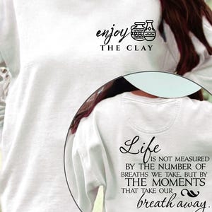 Könnte beinhalten: Weißes Sweatshirt mit dem Text "enjoy the clay" und einer Grafik von zwei Vasen. Auf der Rückseite des Sweatshirts steht der Text "Life is not measured by the number of breaths we take, but by the moments that take our breath away."