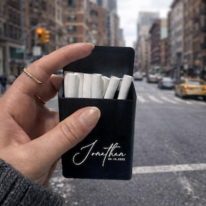Puede incluir: Un estuche negro para cigarrillos con el nombre "Jonathan" y la fecha "06.16.2022" en escritura blanca. El estuche está abierto, revelando varios cigarrillos blancos. El fondo es una calle de la ciudad con coches y edificios.