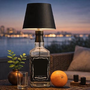 Puede incluir: Una lámpara de mesa única hecha de una botella de whisky reciclada, con una pantalla negra. La lámpara está sobre una mesa de mimbre con una naranja, un pequeño jarrón con vegetación y un vaso. El fondo muestra un horizonte urbano al anochecer.