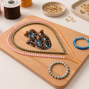 May include: A wooden craft board with a teardrop-shaped cutout is surrounded by strands of beads in pink and green. A blue beaded bracelet and a gray beaded bracelet are also visible. Spools of thread and jewelry-making tools are in the background.