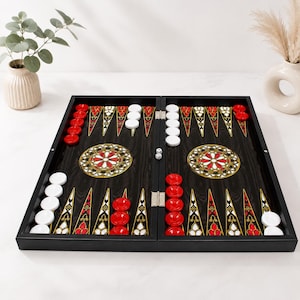 May include: A black backgammon board featuring a dark wood grain finish, decorated with intricate red, white, and gold triangular points. The board includes circular red and white playing pieces, two small dice, and ornate circular center motifs.
