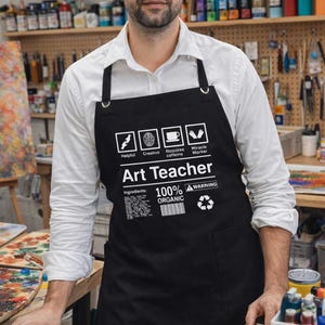 Puede incluir: Delantal negro con el texto "Art Teacher" y las palabras "Helpful, Creative, Requires caffeine, Miracle Worker". El delantal también tiene el texto "100% Organic" y un símbolo de reciclaje. El delantal se lleva sobre una camisa blanca.