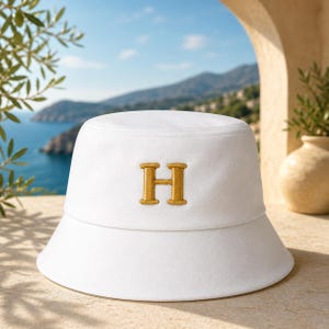 May include: White bucket hat with a gold embroidered 'H' on the front. The hat is made of a soft material and has a wide brim. The background shows a scenic view of the sea and mountains.