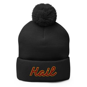 May include: Black beanie hat with a large pom-pom. The word "Hail" is embroidered in red with a gold outline. A winter hat for warmth and style.
