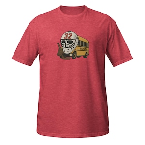 Puede incluir: Camiseta roja con un diseño de dibujos animados. El diseño incluye una máscara de portero de hockey con el número 32, un autobús escolar amarillo con la palabra "BUSSI" y palos de hockey. La camiseta es de un material suave.