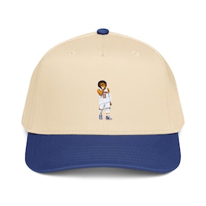 Puede incluir: Gorra de béisbol con corona color crema y visera azul real. La gorra presenta un pequeño gráfico de un jugador de baloncesto con una camiseta blanca con el número 11. La gorra tiene un botón azul en la parte superior.