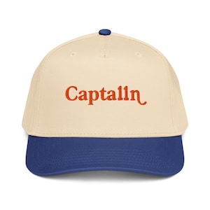 Puede incluir: Gorra de béisbol con corona color crema y visera azul. La palabra "Captalln" está bordada en naranja en la parte delantera de la gorra. La gorra tiene un botón azul en la parte superior.
