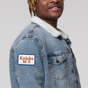 May include: A light blue denim jacket with a white sherpa collar and a rectangular patch on the sleeve. The patch has a white background with a blue border and features the orange text "Knicks in 5" printed in a bold, serif font style.