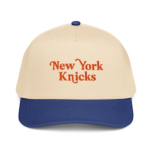 Puede incluir: Gorra de béisbol con corona color crema y visera azul. La gorra presenta el texto "New York Knicks" en escritura naranja en la parte delantera. La gorra tiene un botón azul en la parte superior.