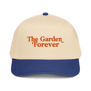 Puede incluir: Gorra de béisbol con corona color crema y visera azul marino. La gorra presenta el texto "The Garden Forever" en letras naranjas en la parte delantera. La gorra tiene un botón azul marino en la parte superior.