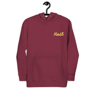 May include: A maroon hoodie with a kangaroo pocket and a drawstring hood. The word "Hail" is embroidered in yellow script on the upper chest. The hoodie is hanging on a black metal hanger.