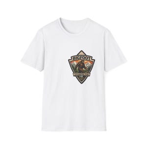 May include: White t-shirt featuring a Bigfoot Exploration Team graphic. The graphic is a triangular patch with a brown Bigfoot figure in a mountain landscape, with the words "BIGFOOT" and "EXPLORATION TEAM".