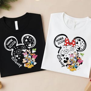 two t-shirts with Disney-themed designs, including Mickey Mouse and Minnie Mouse characters. The t-shirts are placed on a surface with Christmas decorations, such as ornaments and a Christmas tree.