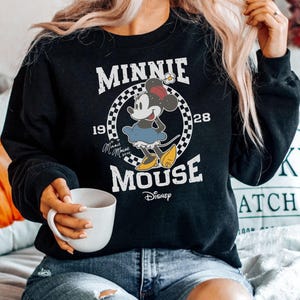 A woman wearing a black sweatshirt with a Minnie Mouse graphic and holding a white mug.