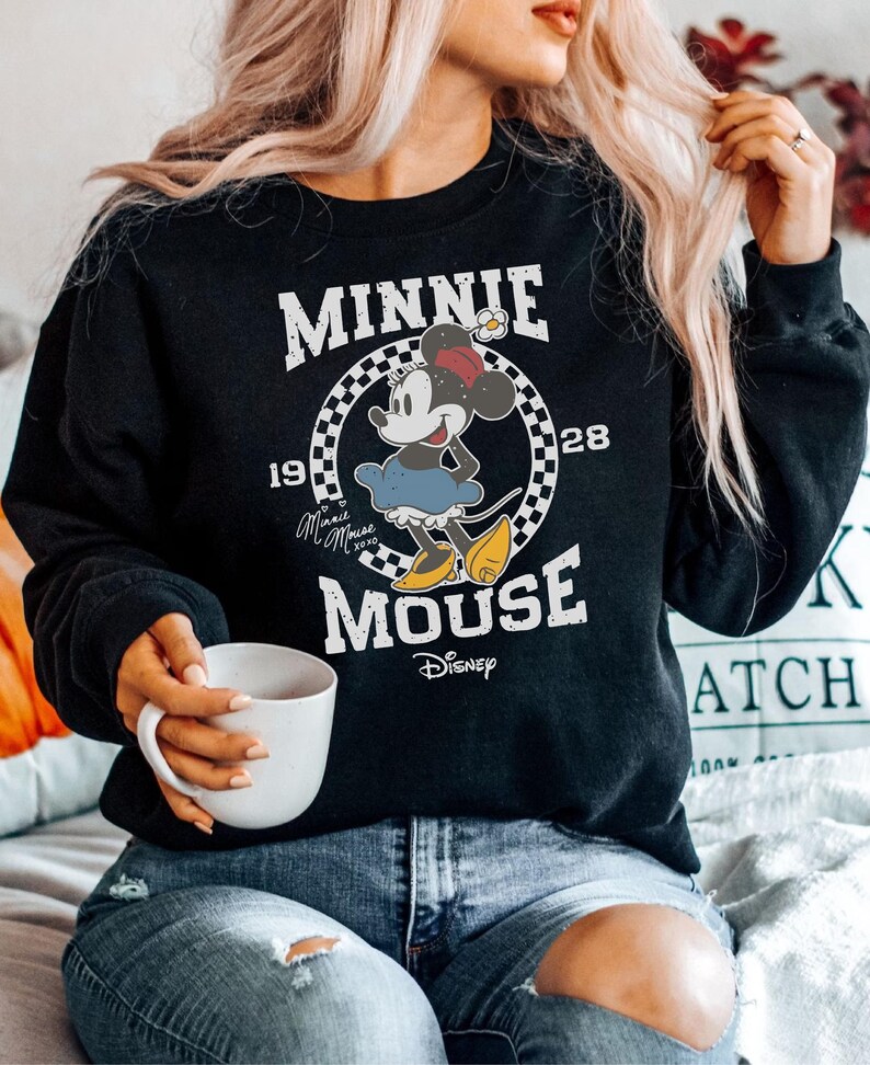 A woman wearing a black sweatshirt with a Minnie Mouse graphic and holding a white mug.