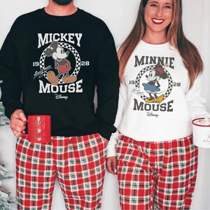 A man and a woman are standing next to each other, both wearing Mickey Mouse-themed pajamas. The man is holding a red mug, and the woman is holding a white mug. They are both smiling and appear to be enjoying themselves.