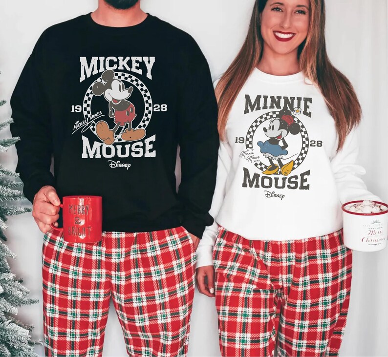 A man and a woman are standing next to each other, both wearing Mickey Mouse-themed pajamas. The man is holding a red mug, and the woman is holding a white mug. They are both smiling and appear to be enjoying themselves.