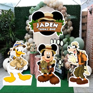Può includere: Decorazioni per feste di compleanno a tema safari con sagome di Topolino, Paperino e Pippo. Le decorazioni includono un cartello con la scritta "Jaden Turns One". Palloncini e una torta completano la scena della festa.