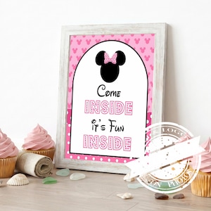 May include: A framed pink and white sign with the text "Come Inside It's Fun Inside" in pink letters. The sign features a black silhouette of Minnie Mouse's head with a pink bow. Cupcakes with pink frosting are in the foreground.