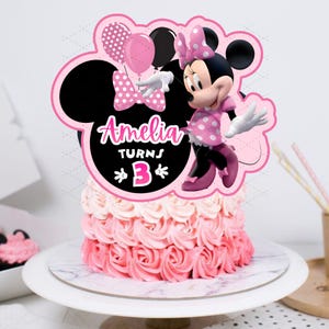 May include: A pink and white ombre cake with a Minnie Mouse cake topper. The topper features Minnie Mouse, balloons, and the text "Amelia Turns 3". The cake is on a white cake stand.