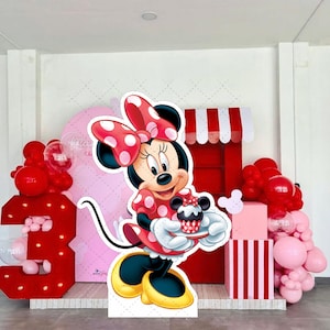 May include: A Minnie Mouse-themed party decoration set. Includes a large cutout of Minnie Mouse holding a cupcake, red and pink balloons, a number 3, and a decorative stand with a red and white striped design. The backdrop features a pink arch.