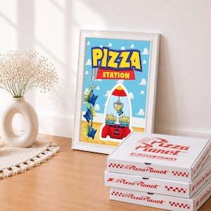 May include: A framed wall art print titled Pizza Station featuring green three-eyed aliens and a red claw machine, displayed next to a stack of three white Pizza Planet cardboard pizza boxes with red text and a checkered border design.