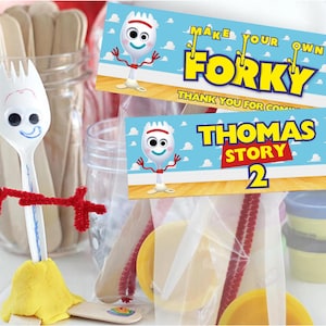 Può includere: Una forchetta di plastica bianca con un volto disegnato e braccia in scovolini rossi e una base gialla. Sono visibili anche due banner blu con il testo "MAKE YOUR OWN FORKY" e "THOMAS STORY 2", insieme a materiali per l'artigianato.