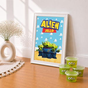 May include: A framed print featuring the text ALIEN JELLO in yellow and red letters against a blue sky background with white clouds, depicting three green three-eyed aliens. Four clear plastic cups of green gelatin with alien faces are nearby.