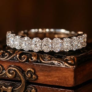 May include: A silver ring with multiple oval-cut diamonds. The ring is displayed on a dark wooden jewelry box with ornate metal detailing. The box is sitting on a cream-colored lace surface.