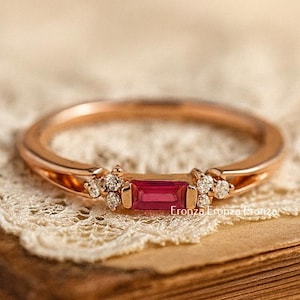 May include: Rose gold ring featuring a rectangular ruby gemstone and small diamond accents. The ring is set on a vintage book with a lace doily. The ring is a piece of jewelry.